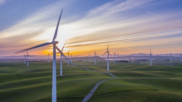 Group of wind turbines on rolling fields, with sun setting in background.