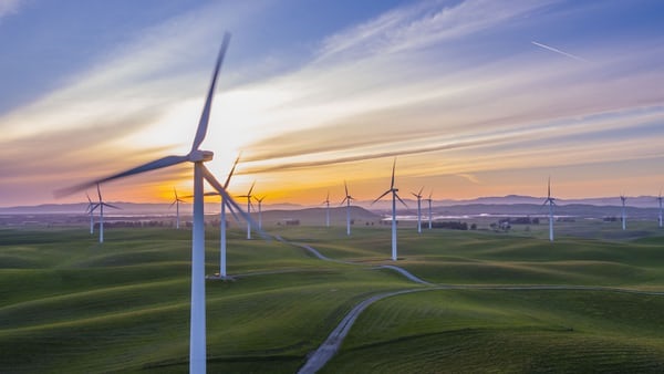 photo-1522735338363-cc7313be0ae0 Group of wind turbines on rolling fields, with sun setting in background.