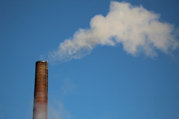 photo-1580064461598-505b080a8242 Pipe blowing out clouds of white smoke into blue sky.